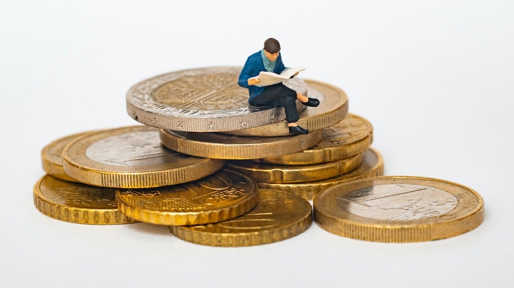 small figurine man reading newspaper on a stack of coins